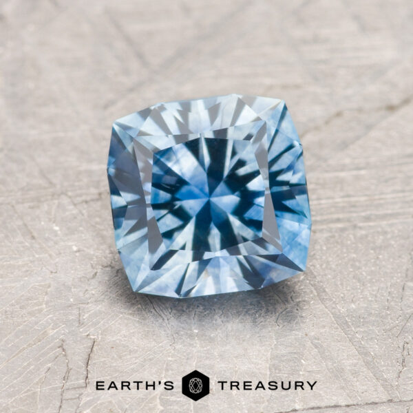 1.52-Carat Sky Blue Montana Sapphire (Heated) A 1.52-carat sky blue Montana Sapphire (heated) with a radiant cut and starburst pattern rests on a textured gray surface, featuring the "Earth’s Treasury" logo below.