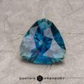 A 1.52-carat deep blue-green Montana Sapphire (heated), faceted in a pear shape, rests on a textured gray surface with the Earth’s Treasury logo and text visible below.