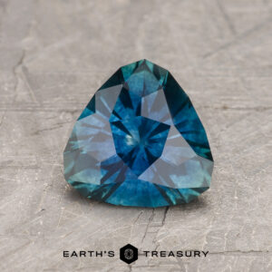 A 1.52-carat deep blue-green Montana Sapphire (heated), faceted in a pear shape, rests on a textured gray surface with the Earth’s Treasury logo and text visible below.