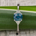 The "Mavis" ring in 14K white gold showcases a 1.02-carat Montana sapphire center and two triangular clear stones, beautifully set against a green leaf background.