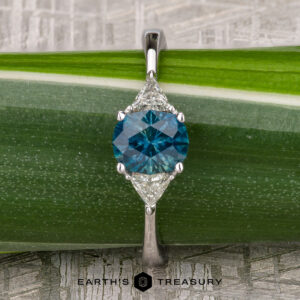 The "Mavis" ring in 14K white gold showcases a 1.02-carat Montana sapphire center and two triangular clear stones, beautifully set against a green leaf background.