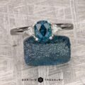 The "Mavis" ring in 14K white gold showcases a 1.02-carat Montana sapphire with two triangular side stones, displayed on a rough blue stone against a textured gray background.