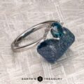The "Mavis" ring in 14K white gold with a 1.02-carat Montana sapphire rests on a rough blue stone, set against a textured silver background. Text at the bottom reads "EARTH'S TREASURY.