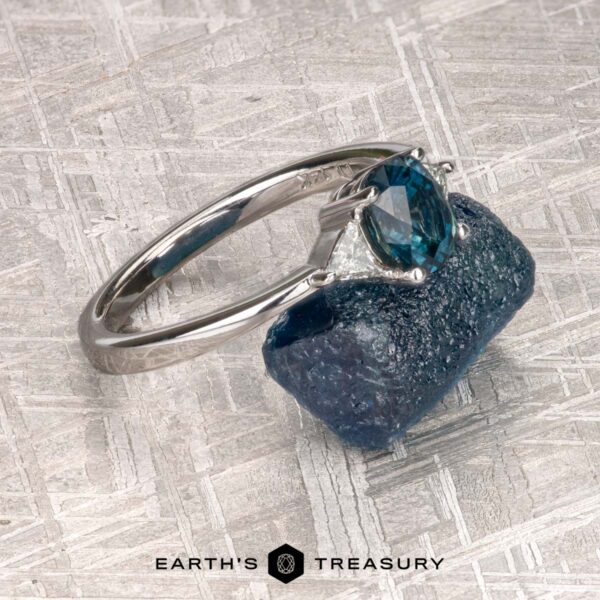 The "Mavis" ring in 14K white gold with 1.02-carat Montana sapphire The "Mavis" ring in 14K white gold with a 1.02-carat Montana sapphire rests on a rough blue stone, set against a textured silver background. Text at the bottom reads "EARTH'S TREASURY.