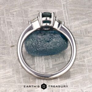 The "Mavis" ring in 14K white gold with a 1.02-carat Montana sapphire is shown upright on a blue stone, with the "Earth's Treasury" logo beneath on a textured background.