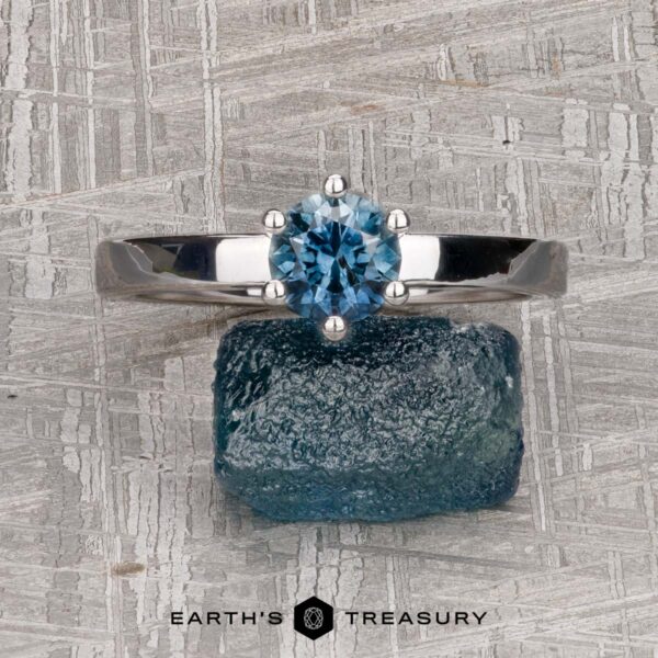 The "Snowflake" ring in 14K white gold, smooth and polished, with 0.95-carat Montana sapphire The "Snowflake" ring in 14K white gold, smooth and polished, with a 0.95-carat Montana sapphire, rests on a rough blue crystal against a textured gray background featuring the "Earth's Treasury" logo at the bottom.