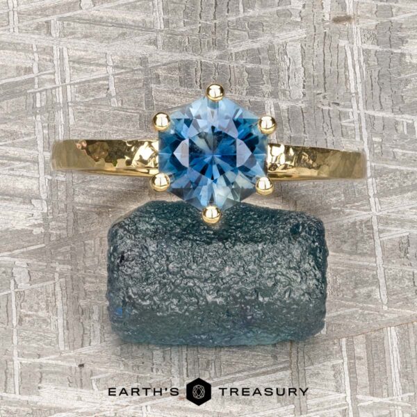 The "Snowflake" ring in 18K yellow gold, hammered and polished, with 1.80-carat Montana sapphire The "Snowflake" ring in 18K yellow gold, hammered and polished, with a 1.80-carat Montana sapphire, is displayed atop a rough blue mineral. "Earth's Treasury" appears at the bottom.