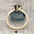 The "Snowflake" ring in 18K yellow gold, hammered and polished, with a 1.80-carat Montana sapphire is displayed upright on a blue rough stone atop a textured surface, with the Earth’s Treasury logo at the bottom.