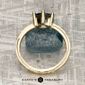 The "Snowflake" ring in 18K yellow gold, hammered and polished, with a 1.80-carat Montana sapphire is displayed upright on a blue rough stone atop a textured surface, with the Earth’s Treasury logo at the bottom.