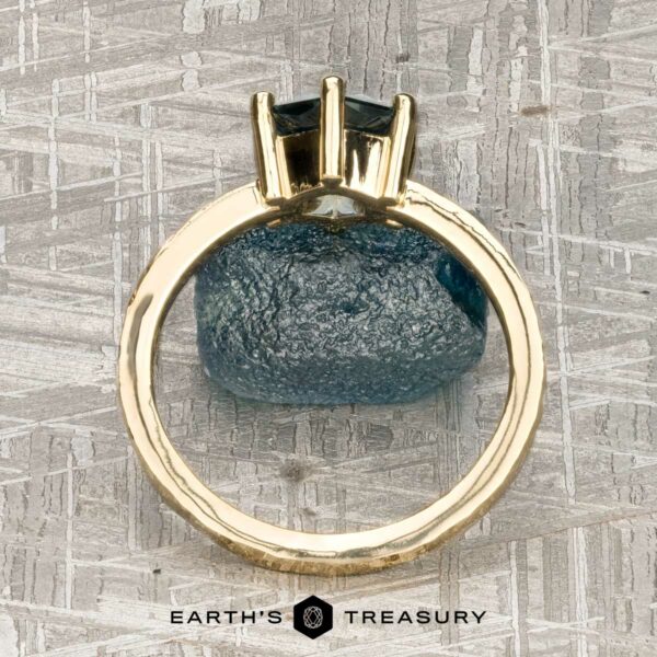 The "Snowflake" ring in 18K yellow gold, hammered and polished, with 1.80-carat Montana sapphire The "Snowflake" ring in 18K yellow gold, hammered and polished, with a 1.80-carat Montana sapphire is displayed upright on a blue rough stone atop a textured surface, with the Earth’s Treasury logo at the bottom.