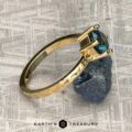 The "Snowflake" ring in 18K yellow gold, hammered and polished, with a 1.80-carat Montana sapphire, stands on a raw blue crystal atop a textured gray surface with "EARTH'S TREASURY" below.