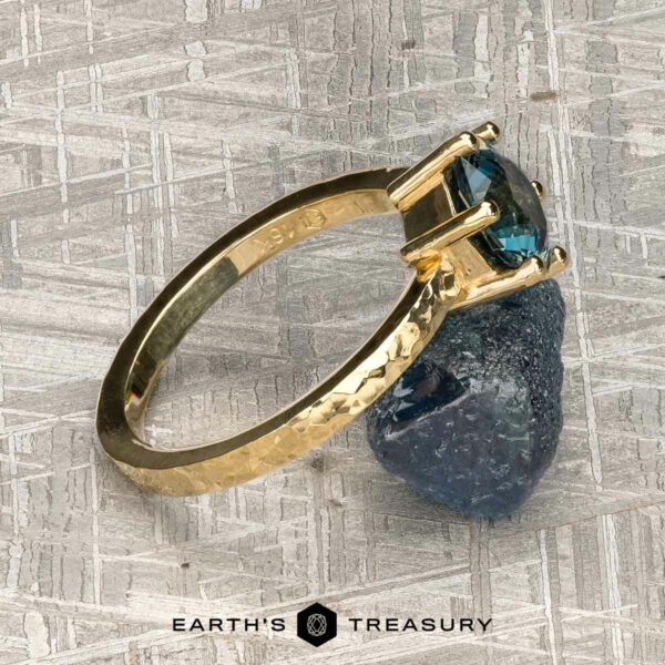 The "Snowflake" ring in 18K yellow gold, hammered and polished, with 1.80-carat Montana sapphire The "Snowflake" ring in 18K yellow gold, hammered and polished, with a 1.80-carat Montana sapphire, stands on a raw blue crystal atop a textured gray surface with "EARTH'S TREASURY" below.