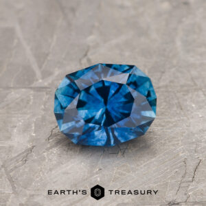 A 1.13-carat Peacock Blue Montana Sapphire (heated) is shown faceted on a gray stone surface, with the Earth's Treasury logo and name featured at the bottom.