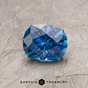 A 1.26-carat rich blue heated Montana Sapphire is shown on a textured gray surface, with "EARTH'S TREASURY" displayed at the bottom.