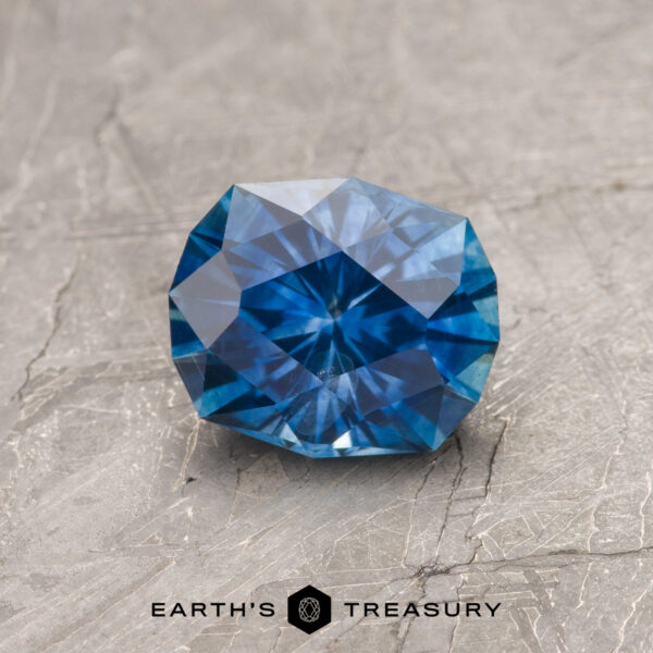 A 1.26-carat rich blue heated Montana Sapphire is shown on a textured gray surface, with "EARTH'S TREASURY" displayed at the bottom.