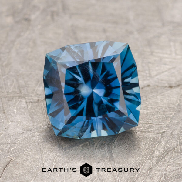 2.40-Carat Rich Blue Montana Sapphire A 2.40-carat Rich Blue Montana Sapphire with cushion cut and star-like facets is displayed on a textured gray surface, with the Earth's Treasury logo at the bottom.