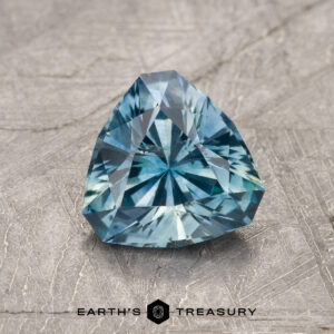 The 1.73-Carat Aqua Blue Montana Sapphire (Heated), trillion-cut and faceted, gleams on a textured gray surface with the Earth’s Treasury logo and name below.