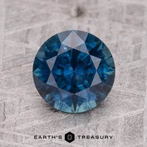 A 2.12-carat faceted Midnight Blue Montana Sapphire (Heated) is displayed on a textured light grey surface with “EARTH’S TREASURY” printed below.