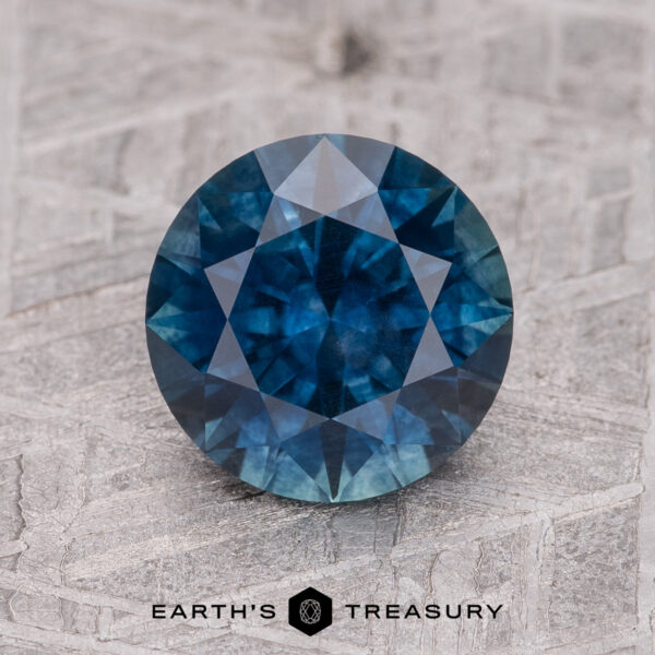 2.12-Carat Midnight Blue Montana Sapphire (Heated) A 2.12-carat faceted Midnight Blue Montana Sapphire (Heated) is displayed on a textured light grey surface with “EARTH’S TREASURY” printed below.