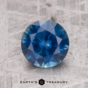 A 1.97-carat rich blue Montana Sapphire (heated), round-cut, sits on a textured gray surface above the "EARTH'S TREASURY" text and geometric logo.