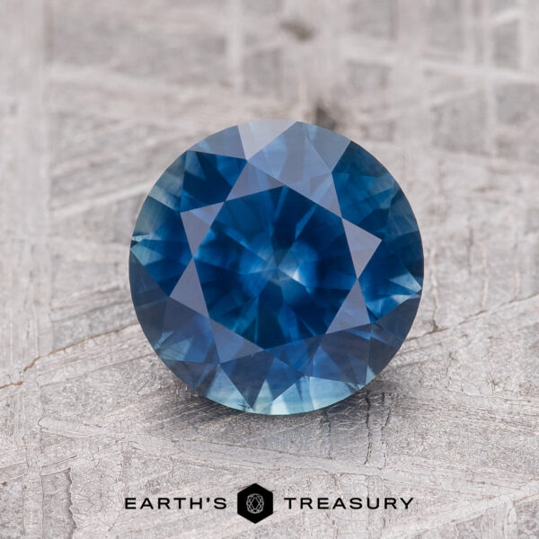 1.97-Carat Rich Blue Montana Sapphire (Heated) A 1.97-carat rich blue Montana Sapphire (heated), round-cut, sits on a textured gray surface above the "EARTH'S TREASURY" text and geometric logo.