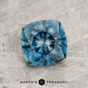 A 1.51-carat teal blue Montana sapphire (heated), cushion-cut with intricate facets, is displayed on a textured gray surface with "Earth's Treasury" branding below.