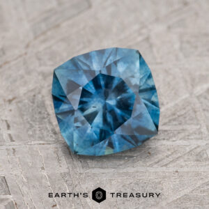 A 1.39-carat medium blue Montana Sapphire (heated), cushion-cut with intricate facets, is displayed on a textured gray surface featuring the text "Earth's Treasury" at the bottom.