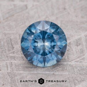 A 1.61-carat teal blue Montana Sapphire (heated), round brilliant-cut, is shown on a textured gray surface with the "Earth's Treasury" logo at the bottom.