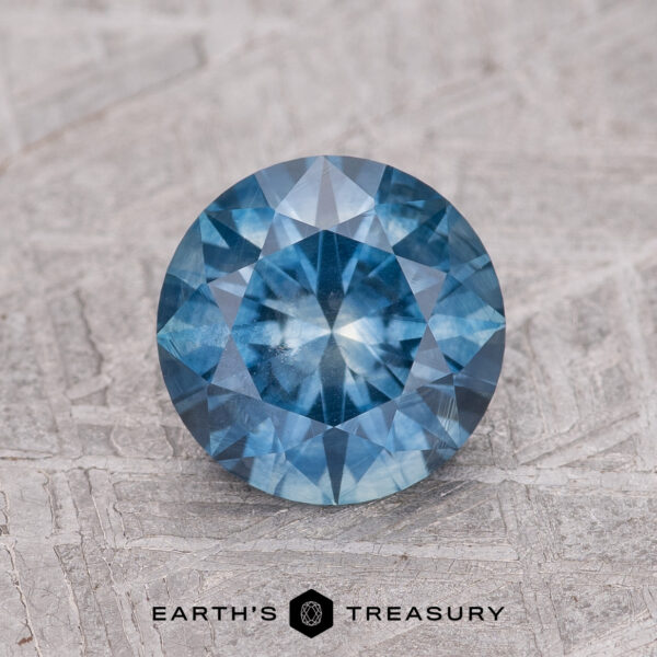 1.61-Carat Teal Blue Montana Sapphire (Heated) A 1.61-carat teal blue Montana Sapphire (heated), round brilliant-cut, is shown on a textured gray surface with the "Earth's Treasury" logo at the bottom.