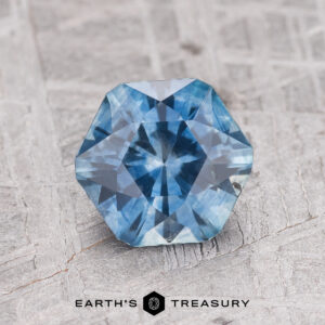 A 1.70-carat Teal Blue Montana Sapphire (Heated) with an octagonal cut is shown on a textured gray surface, with "EARTH'S TREASURY" displayed at the bottom.