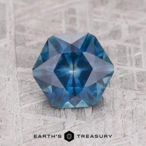 A 1.80-carat dark blue heated Montana sapphire in a hexagon shape with intricate facets, shown on textured gray next to the "Earth's Treasury" logo.