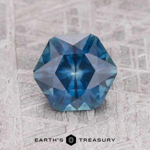 1.80-Carat Dark Blue Montana Sapphire (Heated) A 1.80-carat dark blue heated Montana sapphire in a hexagon shape with intricate facets, shown on textured gray next to the "Earth's Treasury" logo.