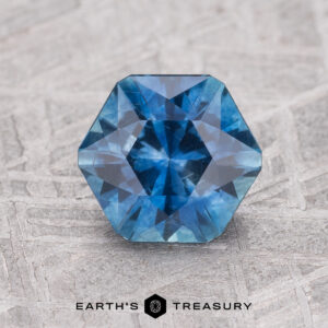 A 1.65-carat rich blue Montana Sapphire (heated), hexagon-cut, gleams against a textured gray surface with the "Earth's Treasury" logo displayed below.