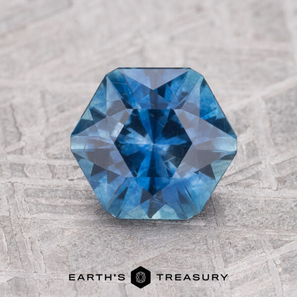 1.65-Carat Rich Blue Montana Sapphire (Heated) A 1.65-carat rich blue Montana Sapphire (heated), hexagon-cut, gleams against a textured gray surface with the "Earth's Treasury" logo displayed below.