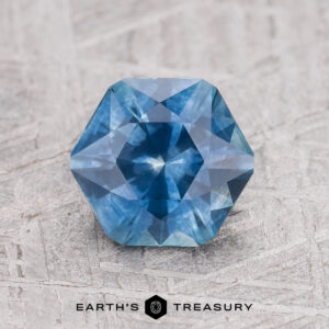 A 1.46-carat medium blue heated Montana Sapphire with a faceted hexagonal shape rests on a textured gray surface featuring the Earth's Treasury logo and name below.