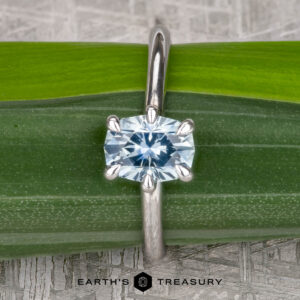 A "Nelisa" solitaire ring in 14K white gold with a 1.51-carat Montana sapphire is displayed on a green leaf. The Earth's Treasury logo is shown at the bottom of the image.