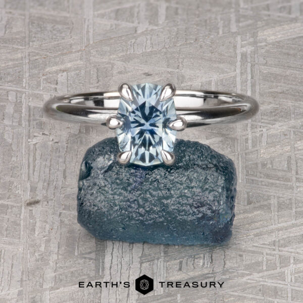 The "Nelisa" ring in 14K white gold with 1.51-carat Montana sapphire The "Nelisa" ring in 14K white gold featuring a 1.51-carat oval-cut Montana sapphire is displayed on a blue stone and set against a textured gray background, with "Earth's Treasury" below.