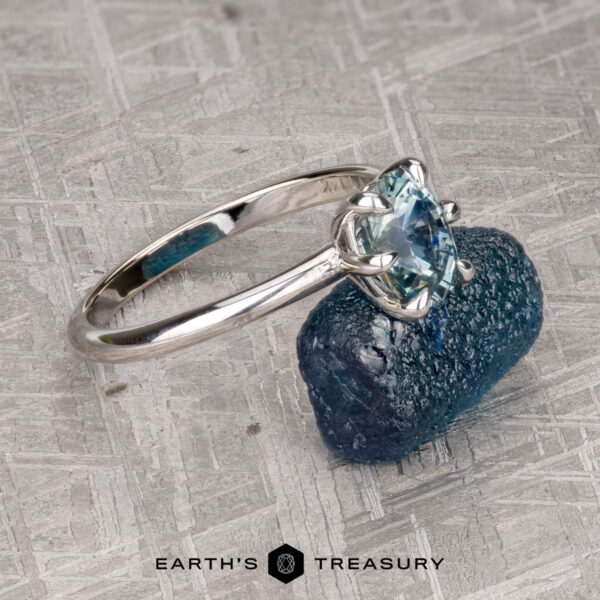 The "Nelisa" ring in 14K white gold with 1.51-carat Montana sapphire The "Nelisa" ring in 14K white gold with a 1.51-carat Montana sapphire rests on a rough blue stone atop a textured gray surface, with Earth's Treasury logo at the bottom.
