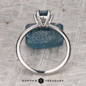 The "Nelisa" ring in 14K white gold with a 1.51-carat Montana sapphire is displayed upright before a rough blue stone on a patterned surface, with "EARTH'S TREASURY" shown at the bottom.