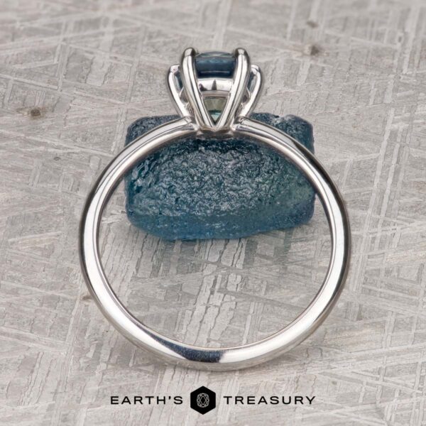 The "Nelisa" ring in 14K white gold with 1.51-carat Montana sapphire The "Nelisa" ring in 14K white gold with a 1.51-carat Montana sapphire is displayed upright before a rough blue stone on a patterned surface, with "EARTH'S TREASURY" shown at the bottom.