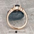 The "Erythia" ring in 20K pink gold, featuring a 1.42-carat Montana sapphire, is shown upright on a rough blue stone with a textured metallic background.