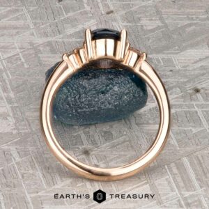 The "Erythia" ring in 20K pink gold, featuring a 1.42-carat Montana sapphire, is shown upright on a rough blue stone with a textured metallic background.