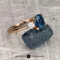 The "Erythia" ring in 20K pink gold, set with a 1.42-carat Montana sapphire, is displayed on a rough blue gemstone atop a textured gray surface. "EARTH'S TREASURY" and Erythia appear below.