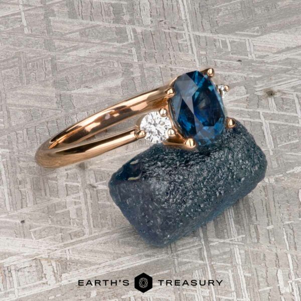 The "Erythia" ring in 20K pink gold with 1.42-carat Montana sapphire The "Erythia" ring in 20K pink gold, set with a 1.42-carat Montana sapphire, is displayed on a rough blue gemstone atop a textured gray surface. "EARTH'S TREASURY" and Erythia appear below.