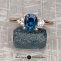 The "Erythia" ring in 20K pink gold with a 1.42-carat Montana sapphire and two diamonds rests on rough blue stone atop a textured gray surface, with "EARTH'S TREASURY" printed below, reflecting the elegance of Erythia jewelry.