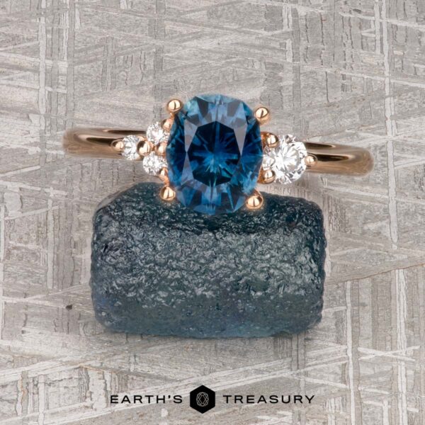 The "Erythia" ring in 20K pink gold with 1.42-carat Montana sapphire The "Erythia" ring in 20K pink gold with a 1.42-carat Montana sapphire and two diamonds rests on rough blue stone atop a textured gray surface, with "EARTH'S TREASURY" printed below, reflecting the elegance of Erythia jewelry.