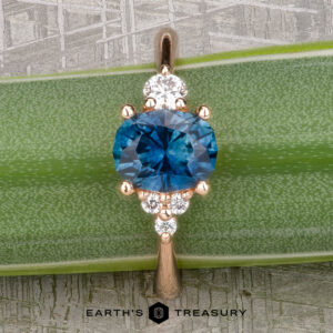 The "Erythia" ring in 20K pink gold, featuring a 1.42-carat oval Montana sapphire and six round diamonds, rests on a green plant leaf above the "EARTH'S TREASURY" text, highlighting exquisite Erythia jewelry.