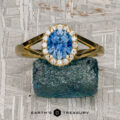 The "Diana" ring in 18K yellow gold, featuring a 1.08-carat Montana sapphire surrounded by small diamonds, rests on a rough blue stone against a textured gray background. "Earth's Treasury" appears at the bottom.