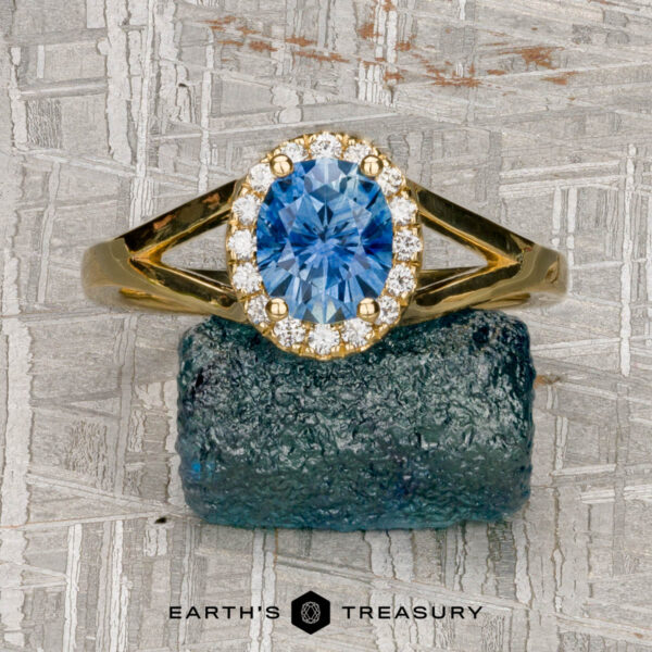 The "Diana" ring in 18K yellow gold with 1.08-carat Montana sapphire The "Diana" ring in 18K yellow gold, featuring a 1.08-carat Montana sapphire surrounded by small diamonds, rests on a rough blue stone against a textured gray background. "Earth's Treasury" appears at the bottom.