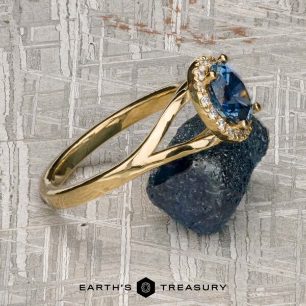 The "Diana" ring in 18K yellow gold with 1.08-carat Montana sapphire The "Diana" ring in 18K yellow gold with a 1.08-carat Montana sapphire, accented by small clear stones, is shown on a rough dark mineral with the Earth's Treasury logo at the bottom.